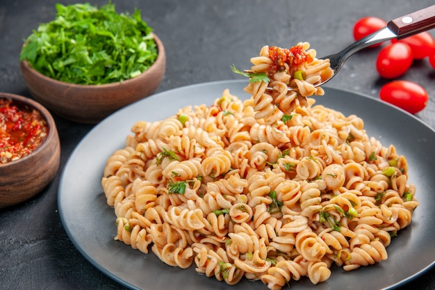 Free photo bottom view rotini pasta on plate on fork cherry tomatoes tomato sauce and chopped greens in bowls on dark isolated surface