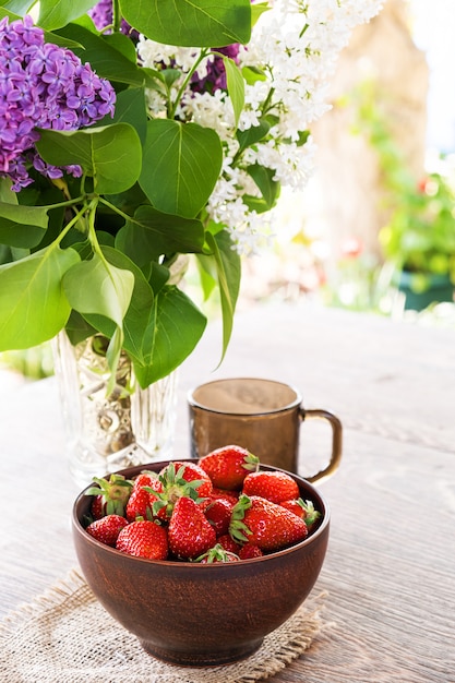 Bouquet of lilac branches in crystal vase, clay bowl with red strawberry and dark glass cup on wooden table.