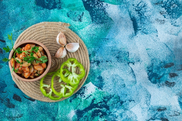 Bowl of baked beans with parsley next to garlic an pepper on a trivet , on the blue table. 