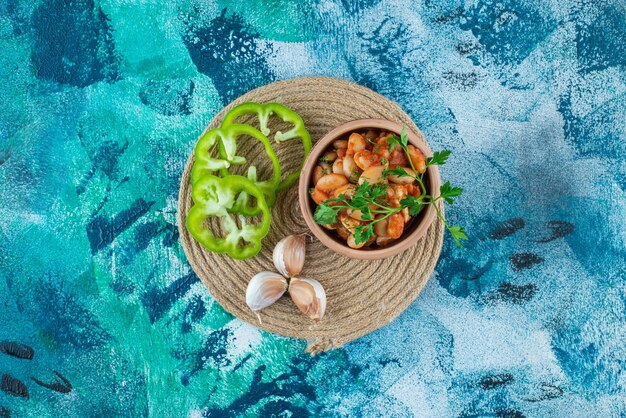 Bowl of baked beans with parsley next to garlic an pepper on trivet on blue.