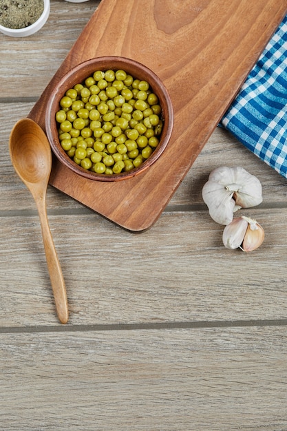 Free Photo a bowl of boiled green peas with a spoon, garlic, and a blue tablecloth on a wooden table.