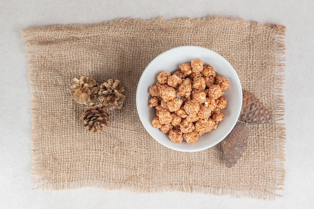 Bowl of caramel popcorn and a bunch of conifer cones on a piece of fabric on marble table.