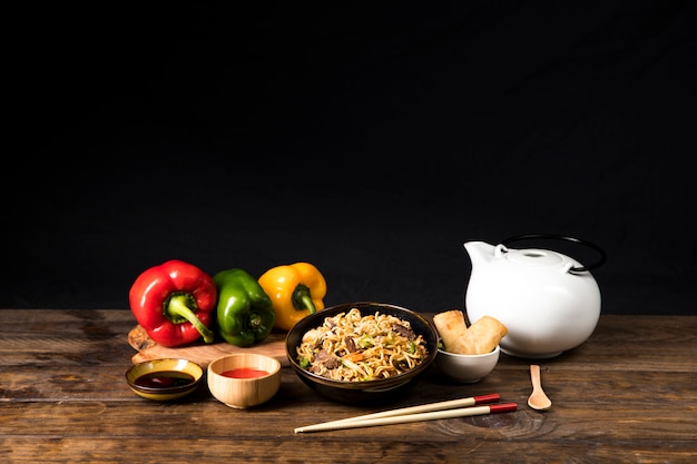 A bowl of delicious beef teriyaki with udon noodles; soya sauce; bell pepper and spring role with chopsticks and spoon on wooden table