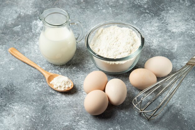 Bowl of flour, eggs and whisker on marble.