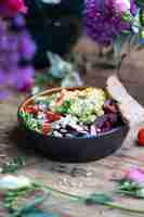 Free photo bowl of healthy salad with avocado, vegetables, and seeds on wooden table among flowers