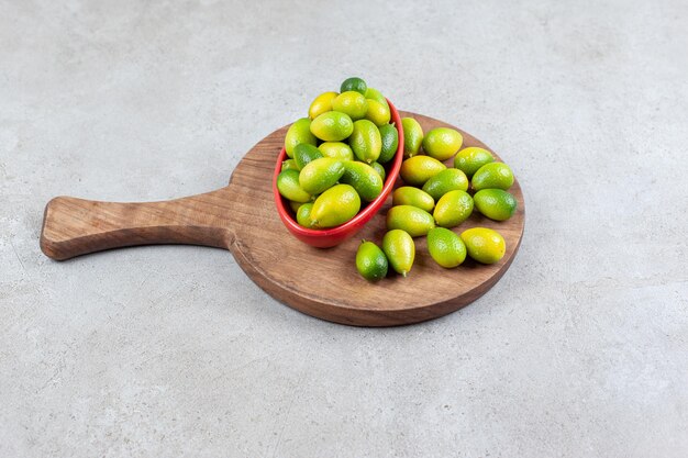 Bowl of kumquats next to a pile on a wooden board in marble surface.