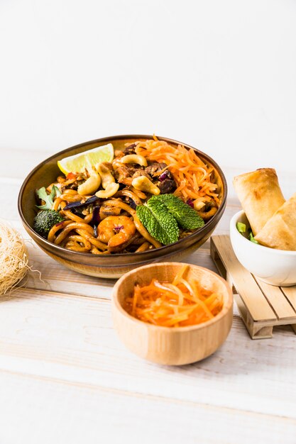 Bowl of noodles with spring roll and grated carrot on wooden table against white backdrop