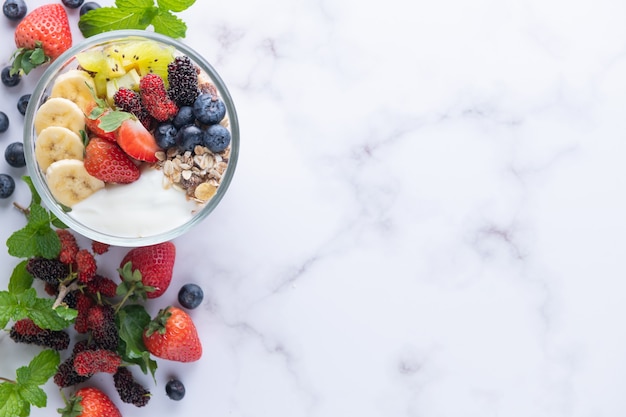 Bowl of oat granola with yogurt, fresh blueberries, mulberry, strawberries, kiwi, banana, mint and nuts board for healthy breakfast, top view, copy space, flat lay. vegetarian food concept.