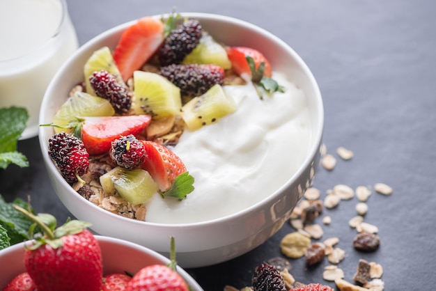 Bowl of oat granola with yogurt, fresh mulberry, strawberries, kiwi mint and nuts on the black rock board for healthy breakfast, copy space. Healthy breakfast menu concept.