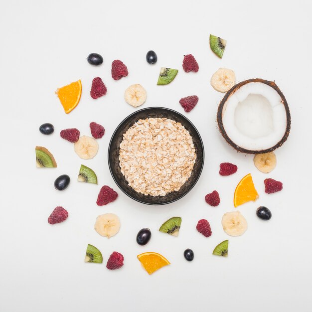 Bowl of oats and halved coconut spread with raspberries; grapes; banana; kiwi slices on white background