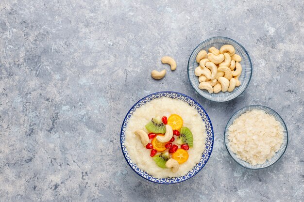 Bowl of rice flakes porridge with kiwi slices, pomegranate seeds, cumquats and cashew nuts, top view