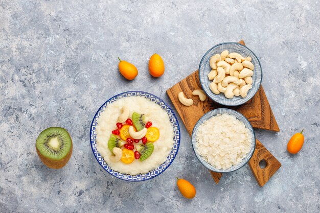 Bowl of rice flakes porridge with kiwi slices, pomegranate seeds, cumquats and cashew nuts, top view