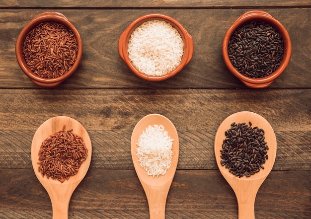 Bowl and wooden spoon with white; red and black rice grains on wooden table