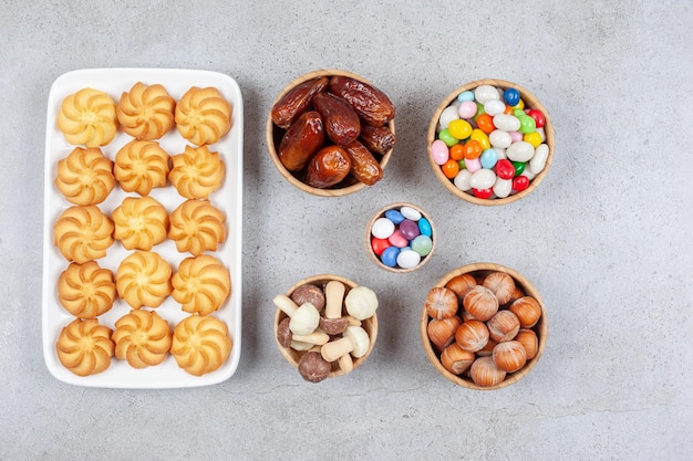 Bowls of candies, hazelnuts, dates and chocolate mushrooms next to cookies on a plate on marble background. High quality photo