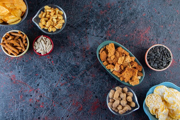 Bowls of crispy chips, crackers and sunflowers seeds on dark surface.