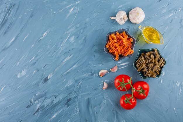 Bowls of multi colored raw spiral pasta with fresh red tomatoes and garlics .