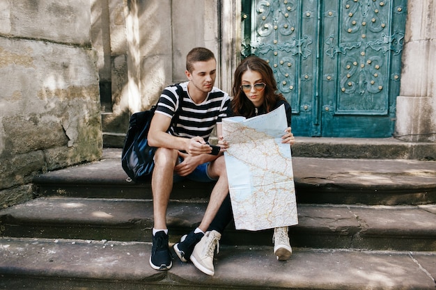 Boy and girl sit on the steps to cathedral with touristc map and watch something in the smartphone