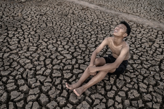 The boy sit hugging their knees bent and looking at the sky to ask for rain on dry soil.