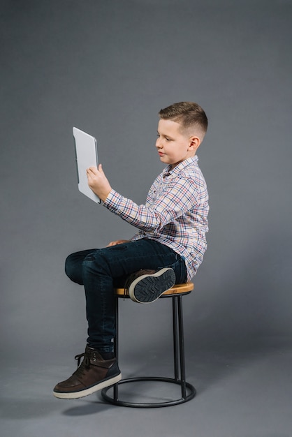 A boy sitting on stool looking at digital tablet against gray background