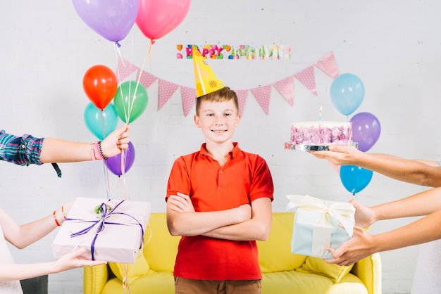 Boy standing between his friend's hand holding birthday cake; gifts and balloons