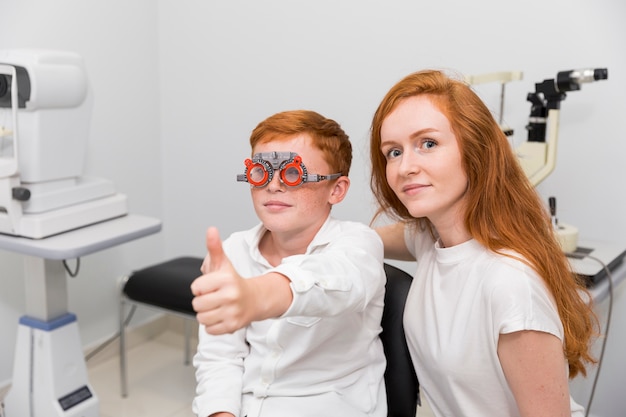 Free Photo boy with optometrist trial frame showing thumb up gesture sitting with young female ophthalmologist in clinic