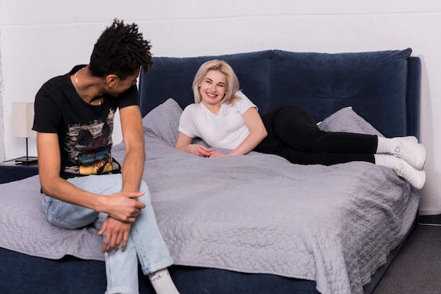 Boyfriend sitting on bed looking at her smiling blonde young woman lying on bed