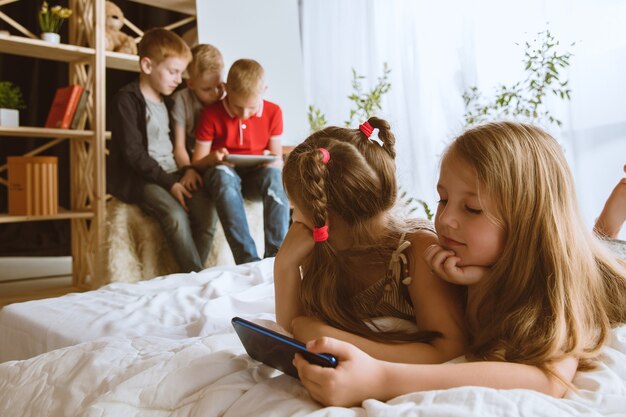 Boys and girls using different gadgets at home. Childs with smart watches, smartphone and headphones. Making selfie, chating, gaming, watching videos. Interaction of kids and modern technologies.