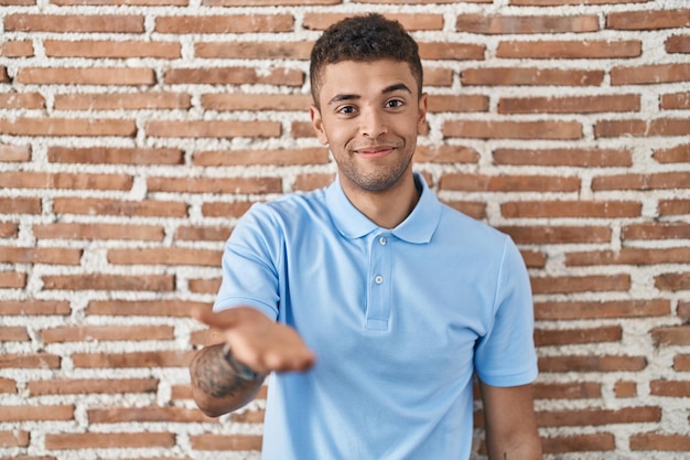 Free photo brazilian young man standing over brick wall smiling cheerful offering palm hand giving assistance and acceptance