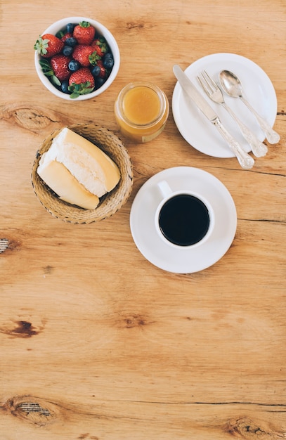 Bread; coffee cup; jam; fresh berries and cutlery on plate against wooden background