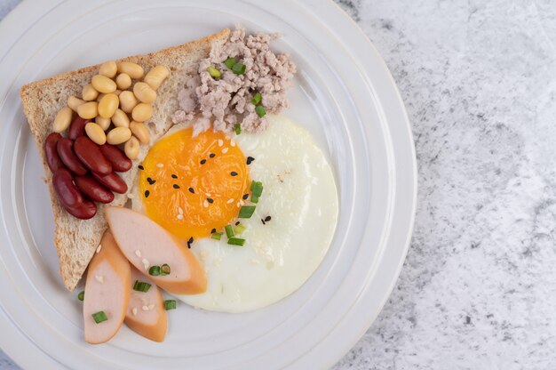 Breakfast consists of fried eggs, sausage, minced pork, bread, red beans and soy on a white plate.