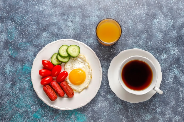 A breakfast plate containing cocktail sausages,fried eggs,cherry tomatoes,sweets,fruits and a glass of peach juice.