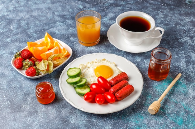 A breakfast plate containing cocktail sausages,fried eggs,cherry tomatoes,sweets,fruits and a glass of peach juice.