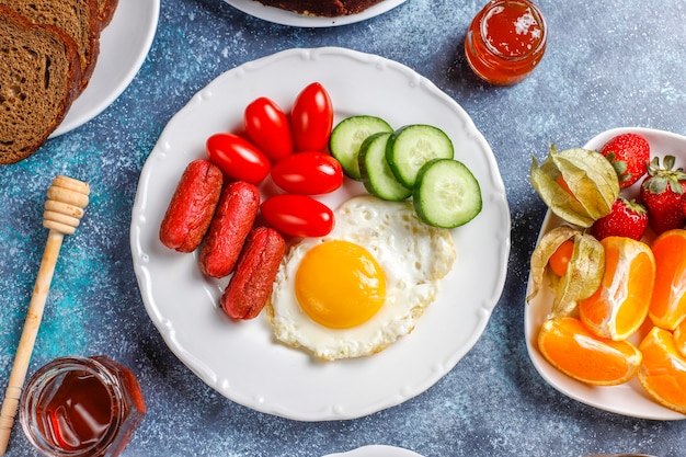 A breakfast plate containing cocktail sausages,fried eggs,cherry tomatoes,sweets,fruits and a glass of peach juice.