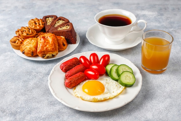 A breakfast plate containing cocktail sausages,fried eggs,cherry tomatoes,sweets,fruits and a glass of peach juice.