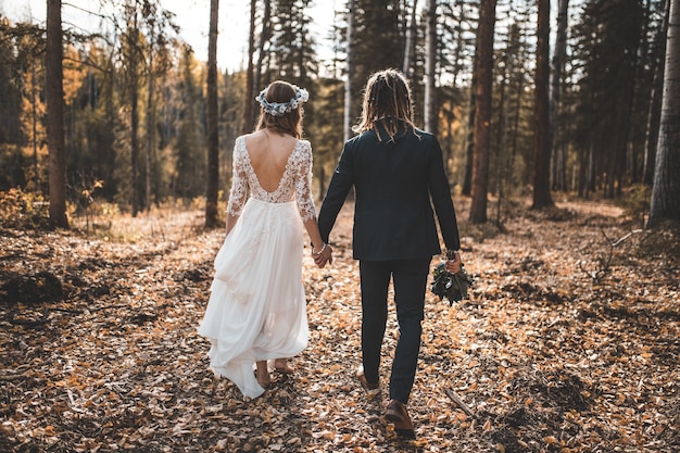 Bride and groom walking on leaves covered ground in woods during day