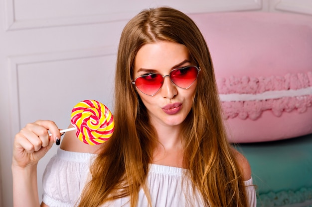 Free photo bright cute portrait of pretty blonde woman holding sweet bright lollipop, long blonde hairs, sugar sweets, hearted sunglasses.