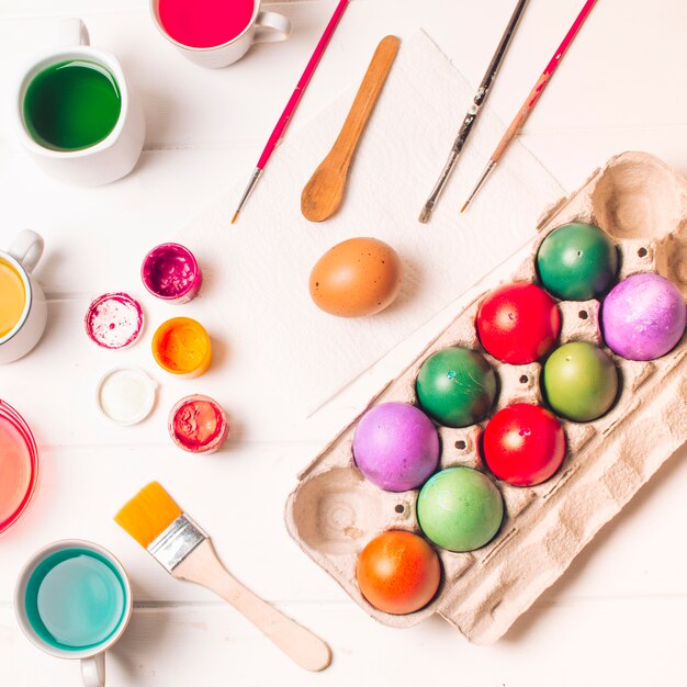 Bright Easter eggs in container near brushes and dye liquid in cups