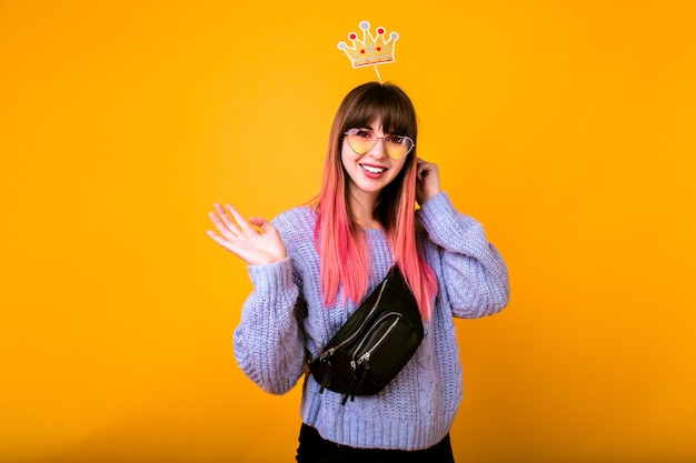 Free photo bright funny portrait of cheerful hipster woman with bright pink hair, wearing cozy sweater, holding fake party crown and smiling, ready for party, yellow wall.