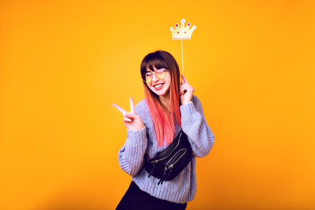 Free photo bright funny portrait of cheerful hipster woman with bright pink hair, wearing cozy sweater, holding fake party crown and smiling, ready for party, yellow wall.