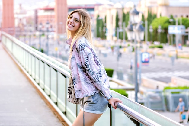 Bright positive sunny spring portrait of happy blonde woman posing at Barcelona square, wearing hipster trendy sportive clothes