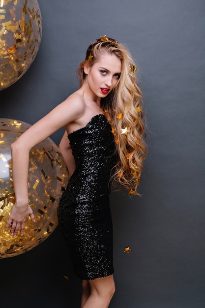 Brightful party time charming young woman in black luxury dress, long curly blonde hair with golden tinsels. Big balloons, party time, fashionable model, cheerful mood.