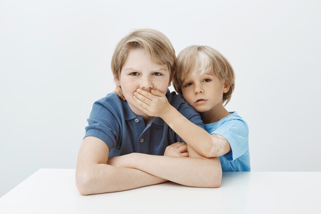 Brother asking to keep secret. Portrait of unhappy annoyed boy sitting at table with hands crossed, frowning while sibling covering his mouth with palm