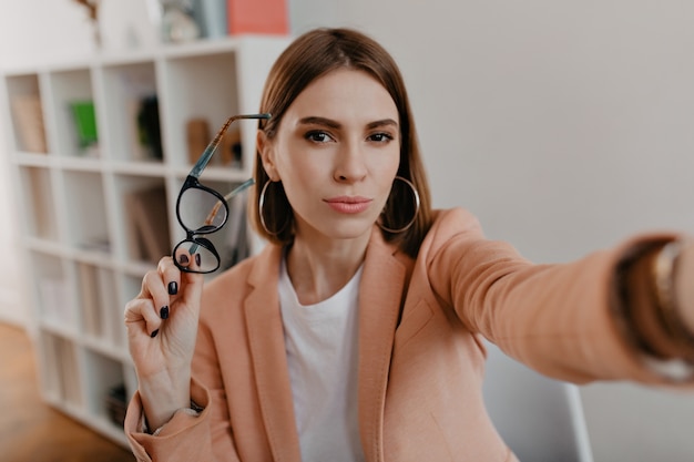 Free photo brown-eyed business woman took off her glasses and takes selfie in her white office.