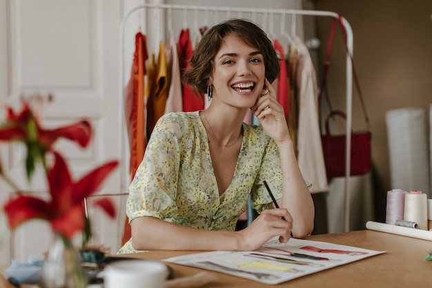 Free photo brown-eyed curly brunette short-haired woman in floral trendy dress smiles, looks at camera, hoods pencil and designs new clothes