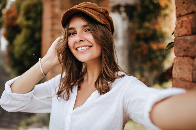 Brown-eyed girl in velvet cap and white blouse makes selfie on space of brick wall and trees.