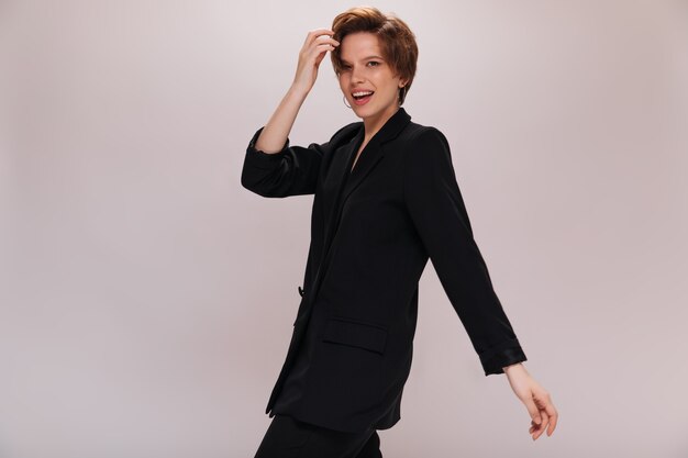 Brown eyed lady in dark suit moves on isolated background. Cool young woman in black jacket and pants walks and smiles on white backdrop