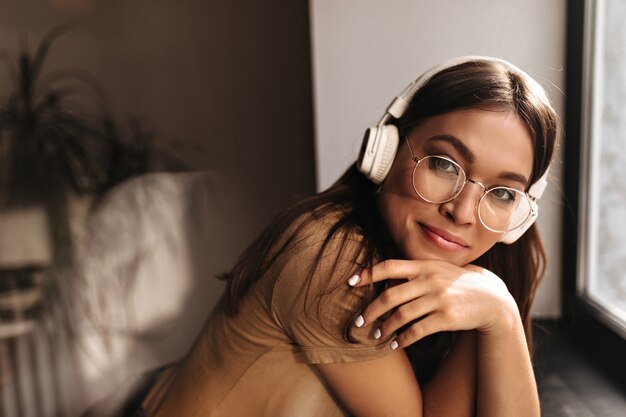 Brown-eyed lady in glasses looking at camera. Portrait of dark-haired woman in white headphones against window.