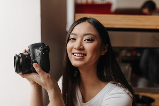 Brown-eyed woman in white top smiles and holds front while posing in her apartment