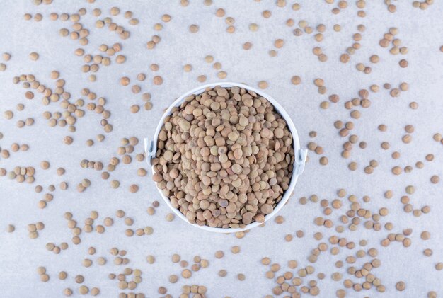 Brown lentil filled into a small bucket sitting in the midst of scattered grains on marble surface