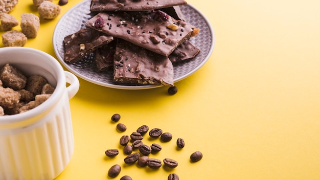 Free Photo brown sugar bowl; roasted coffee beans and chocolate bars on yellow backdrop